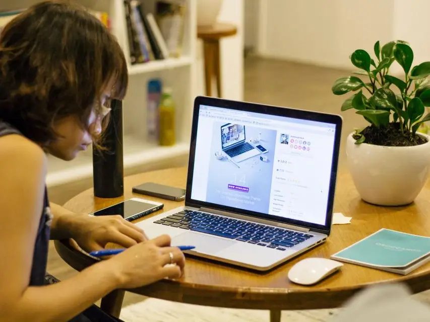 A woman working at a laptop at a round table, a notebook beside her